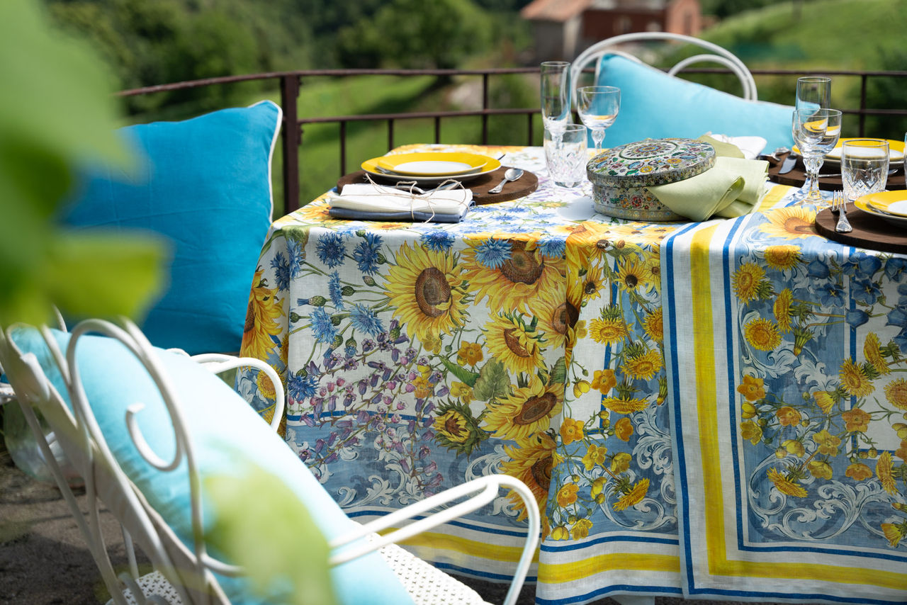 Toalha de Mesa 100% Linho SUNGARDEN- Tessitura Mesa de exterior com toalha estampada de girassóis e cadeiras com almofadas azuis