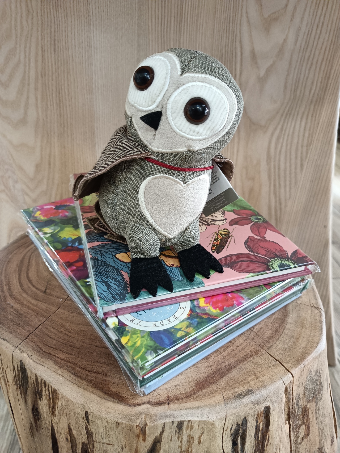 Pisa-Papéis Barn Owl Junior - Mocho Pelúcia de coruja cinzenta com peito em forma de coração em cima de capas florais num tronco de madeira