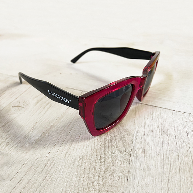 BADDYBOY - ÓCULOS DE SOL TAORMINA Óculos de sol vermelhos e pretos com texto branco na haste, sobre superfície de madeira clara