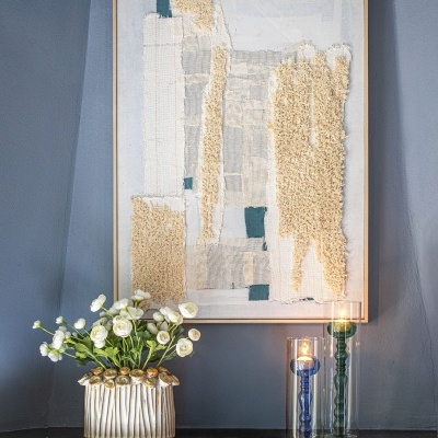 Quadro texturizado de tecidos naturais em tons creme e azul na parede cinzenta, com jarro de flores brancas e castiçais de vidro sobre lareira.