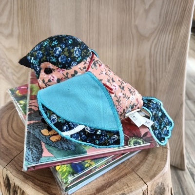 Boneco de tecido em forma de pássaro com padrão floral azul e rosa, sobre livros e tronco de madeira