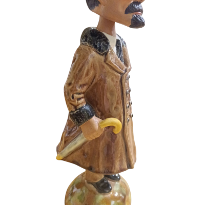 Estatueta cerâmica de homem com casaco castanho e chapéu, segurando bengala amarela