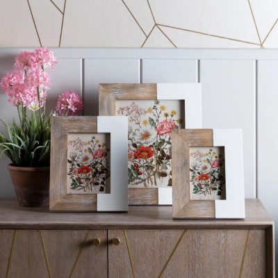 Três molduras de fotos com design dividido madeira e branco e ilustração floral colorida, sobre móvel de madeira com puxadores dourados e flores cor-de-rosa num vaso.