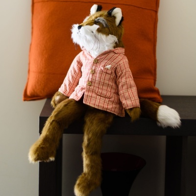 Boneco de peluche de raposa sentado com camisa rosa xadrez e almofada laranja atrás