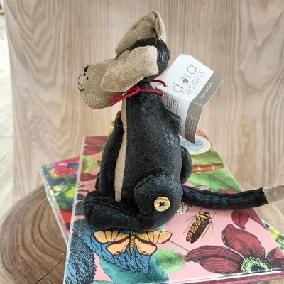 Ratinho de pelúcia preto e bege com laço vermelho e etiqueta, sobre livros florais numa mesa de madeira