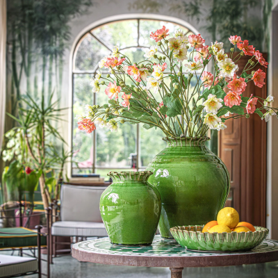 Vasos verdes com flores artificiais sobre mesa redonda com tampo em mosaico num interior com janela arqueada.