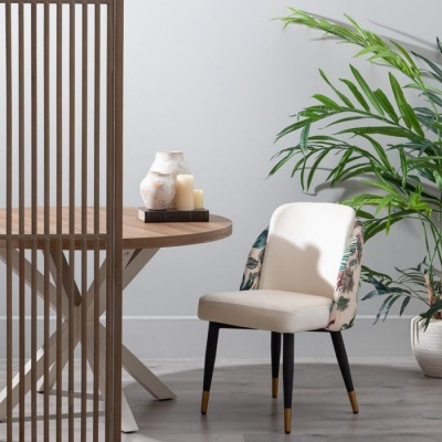 Cadeira estofada creme com padrão floral, mesa de madeira redonda, vela, vaso decorativo e planta verde