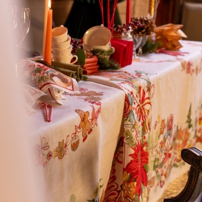 Mesa decorada para Natal com toalha branca e estampas natalinas, velas acesas, chávenas e guardanapos