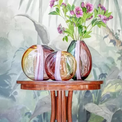 Três vasos coloridos em vidro com flores sobre mesa de madeira e fundo com pintura botânica.