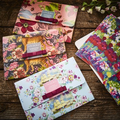 Três envelopes decorativos com estampas florais e animais sobre mesa de madeira e folhas coloridas ao lado.