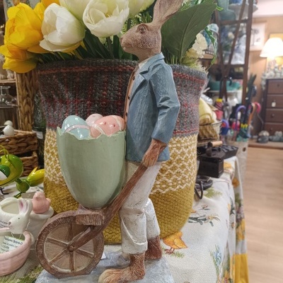 Estatueta de coelho a empurrar carrinho com ovos coloridos, vaso de flores e toalha estampada