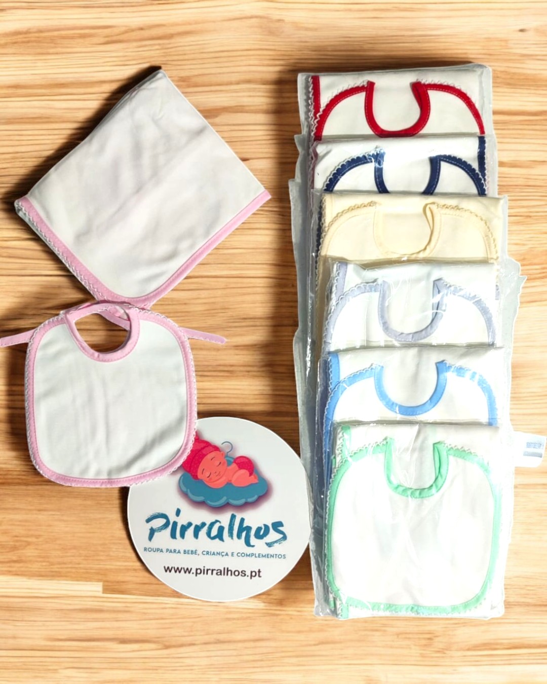 Conjunto de babetes brancos com bordas coloridas e pano branco com borda rosa sobre madeira