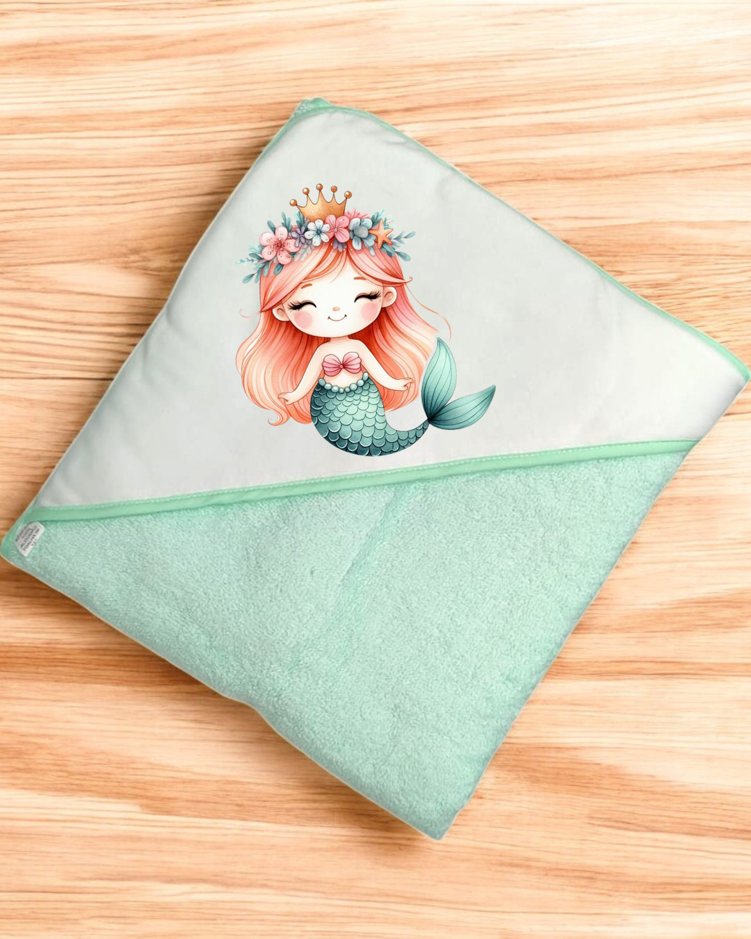 Toalha de bebé verde claro com imagem de sereia e coroa sobre superfície de madeira.