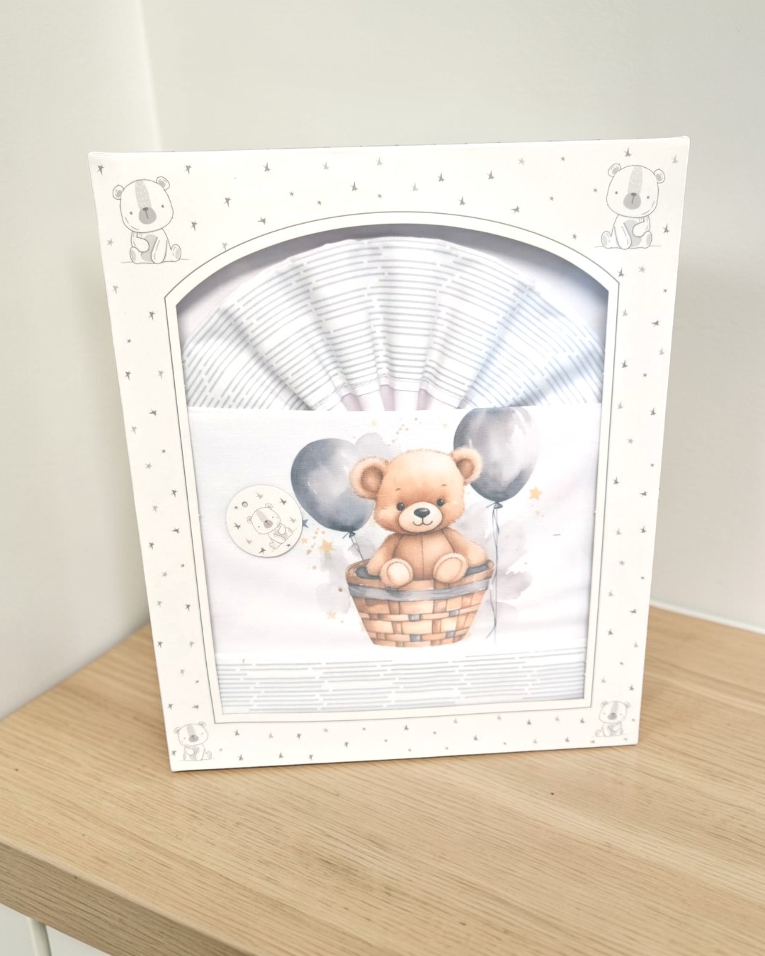 Conjunto de roupa de bebé com urso e balões numa caixa branca decorada com ursos e estrelas.