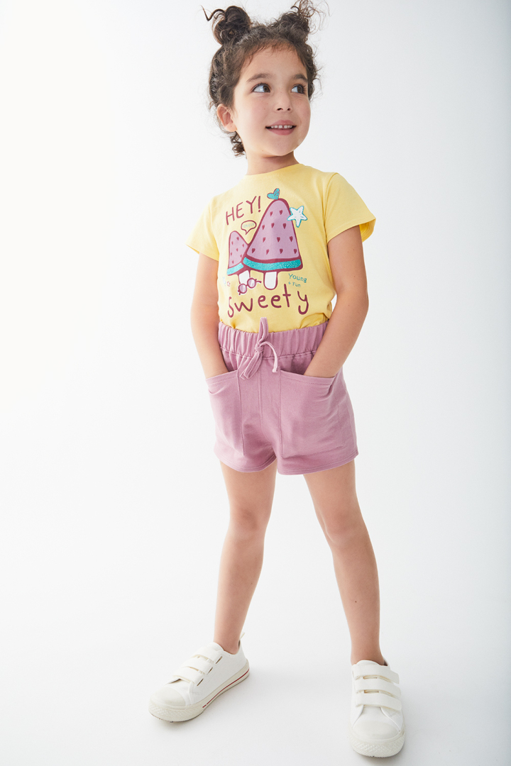 Criança com t-shirt amarela, calções rosa e sapatos brancos em fundo branco