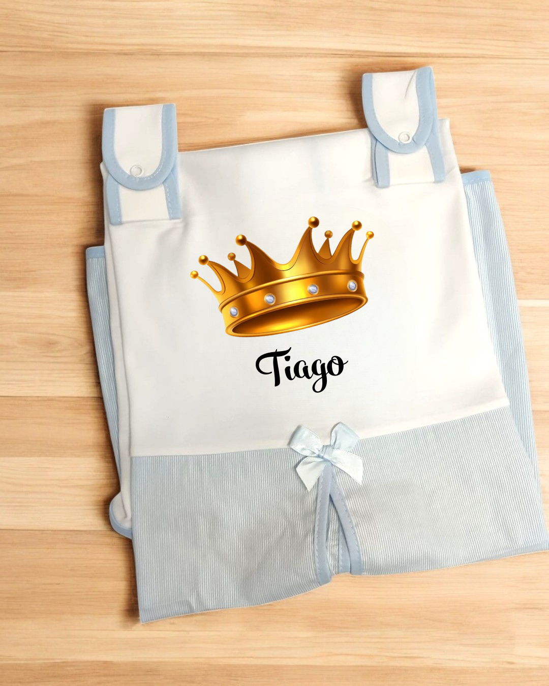 Saco de bebê branco e azul com estampa de coroa dourada e nome Tiago, com laço azul, em superfície de madeira