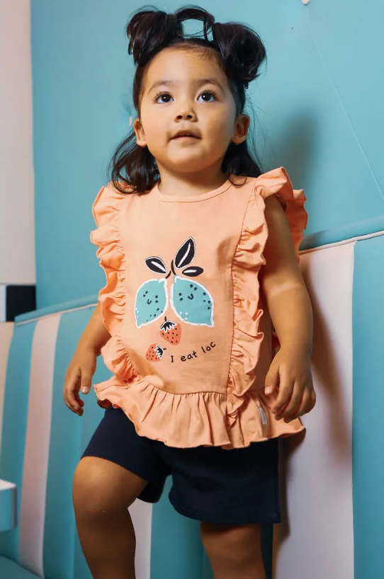 Criança com camisa cor de pêssego com folhos e estampa de folhas e calções escuros