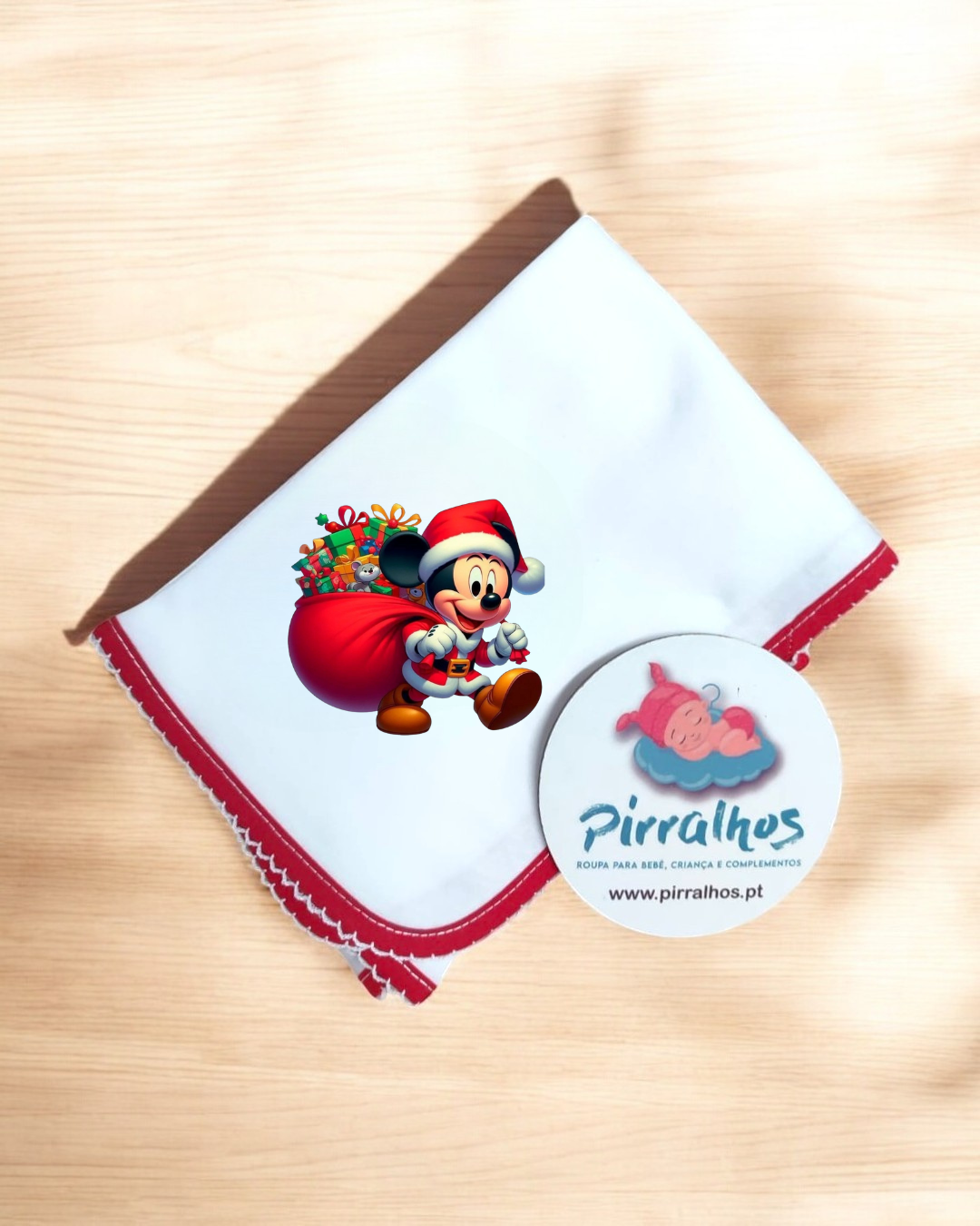 Fralda branca com bordas vermelhas e estampa de Mickey Mouse de Pai Natal com saco de presentes, sobre superfície de madeira clara com etiqueta redonda ao lado.