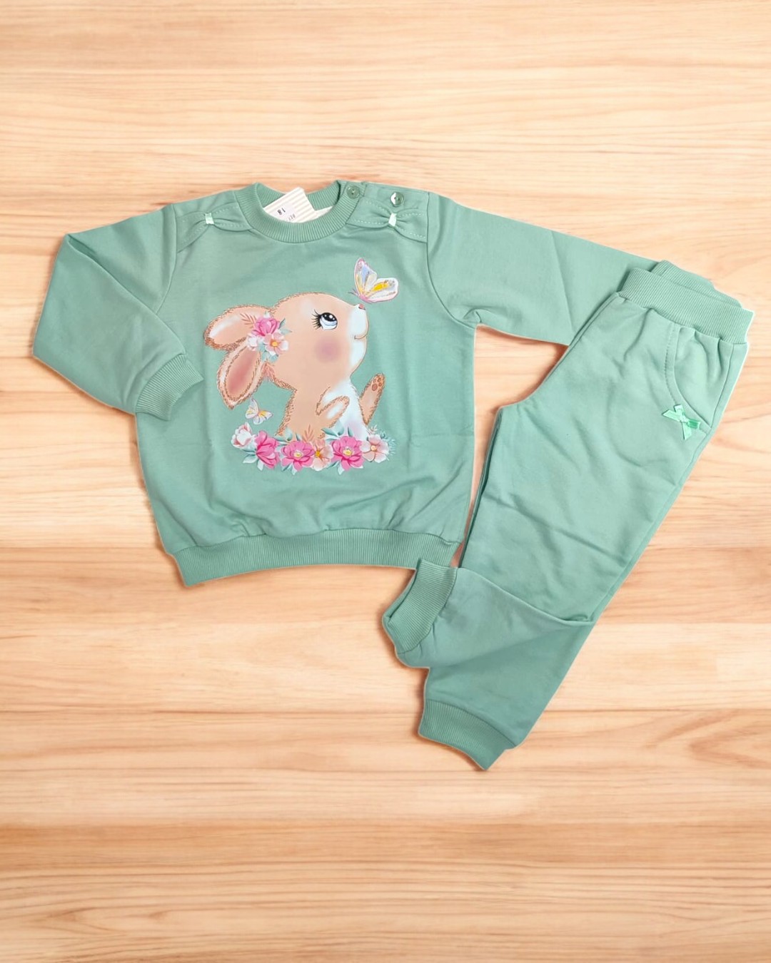 Conjunto infantil verde menta com estampa de coelho e borboleta sobre chão de madeira clara