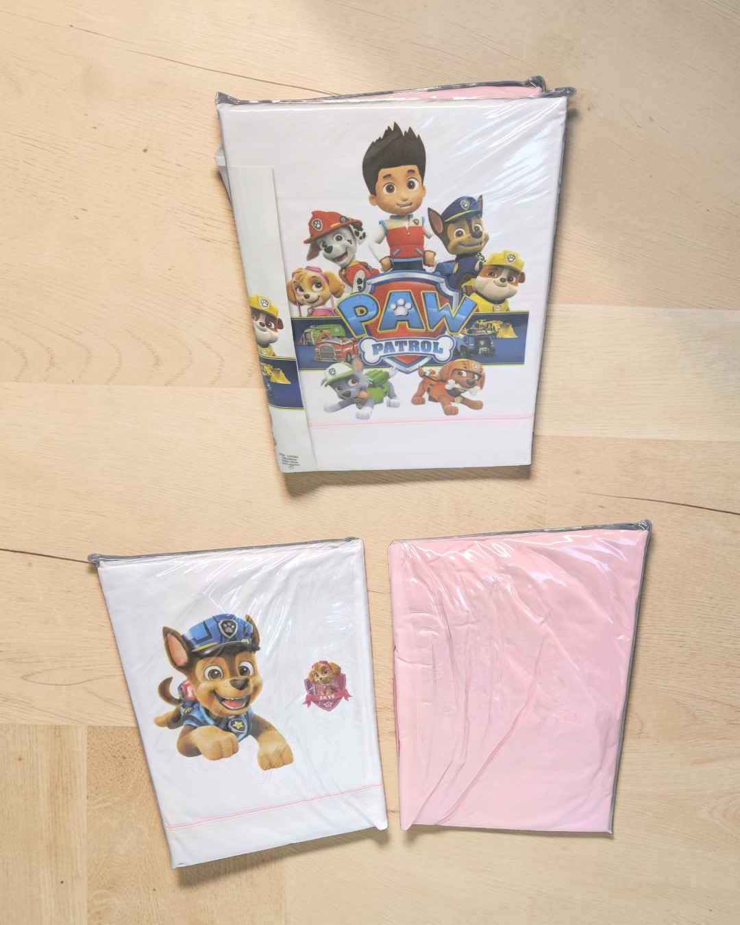 Pacotes de roupa de cama infantil com personagens da Patrulha Pata e tecido rosa sobre chão de madeira.