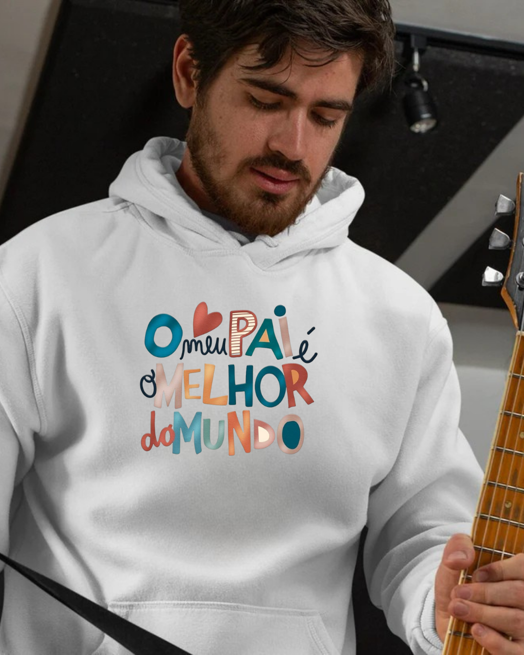 Homem com hoodie branco com texto colorido em português segurando guitarra em interior