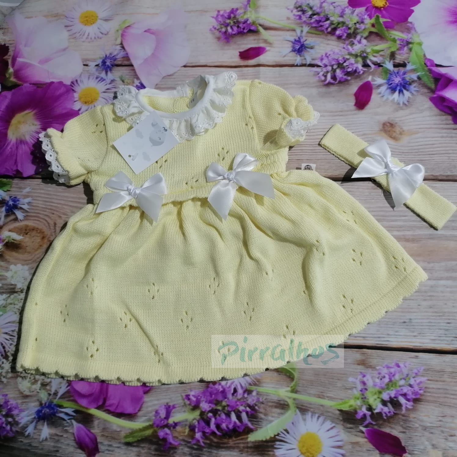 Vestido amarelo claro de malha com detalhes em renda branca e laços, com faixa de cabelo, sobre fundo de madeira com flores