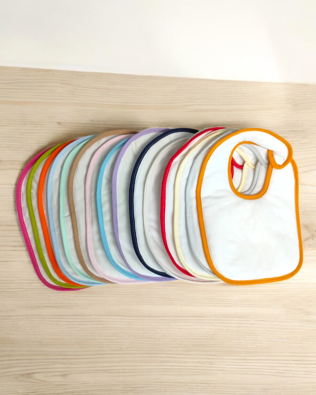 Babetes para bebé brancos com bordas coloridas dispostos em leque sobre madeira clara