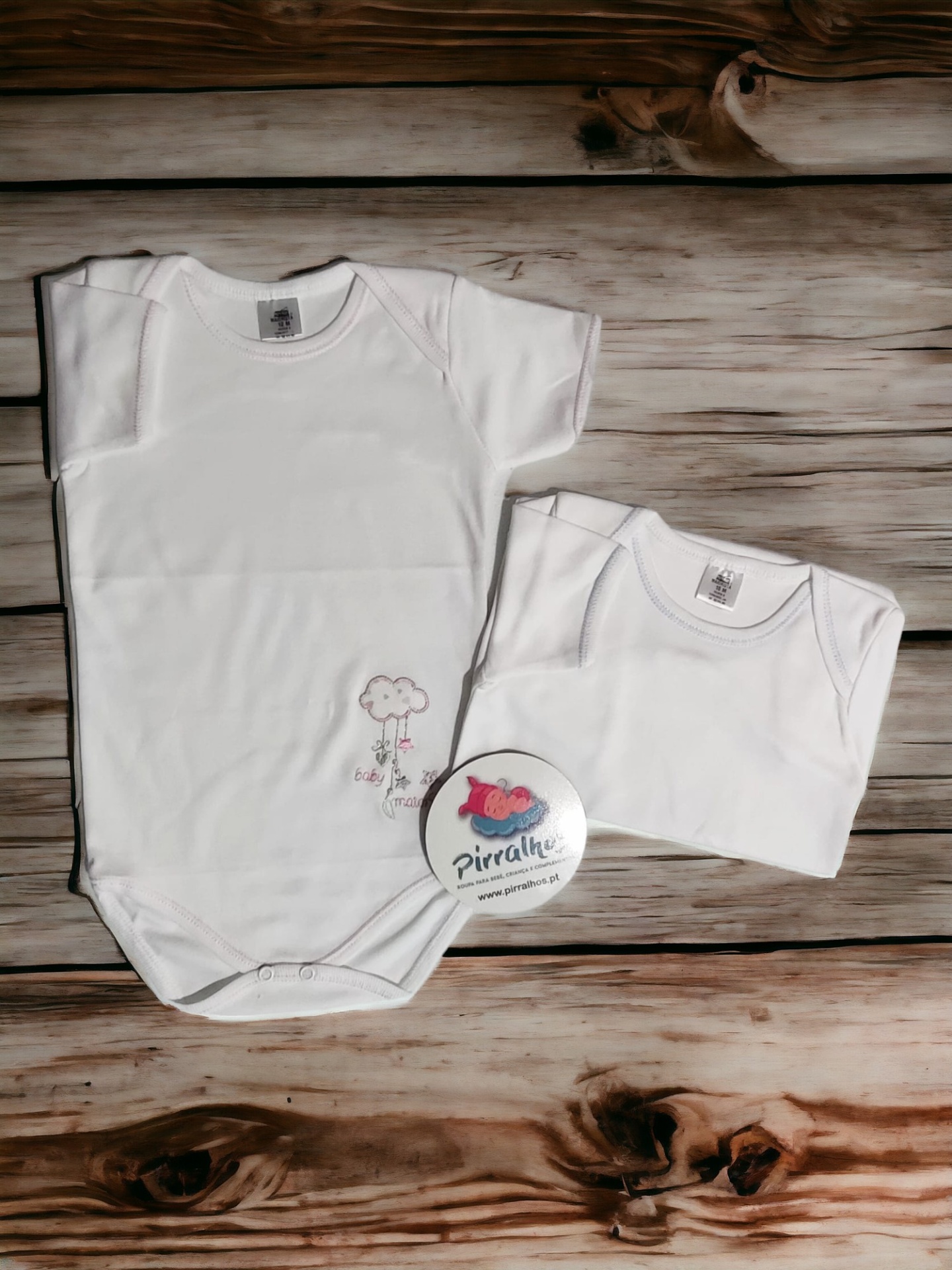 Dois bodies de bebé brancos com um pequeno estampado e um botão promocional sobre madeira