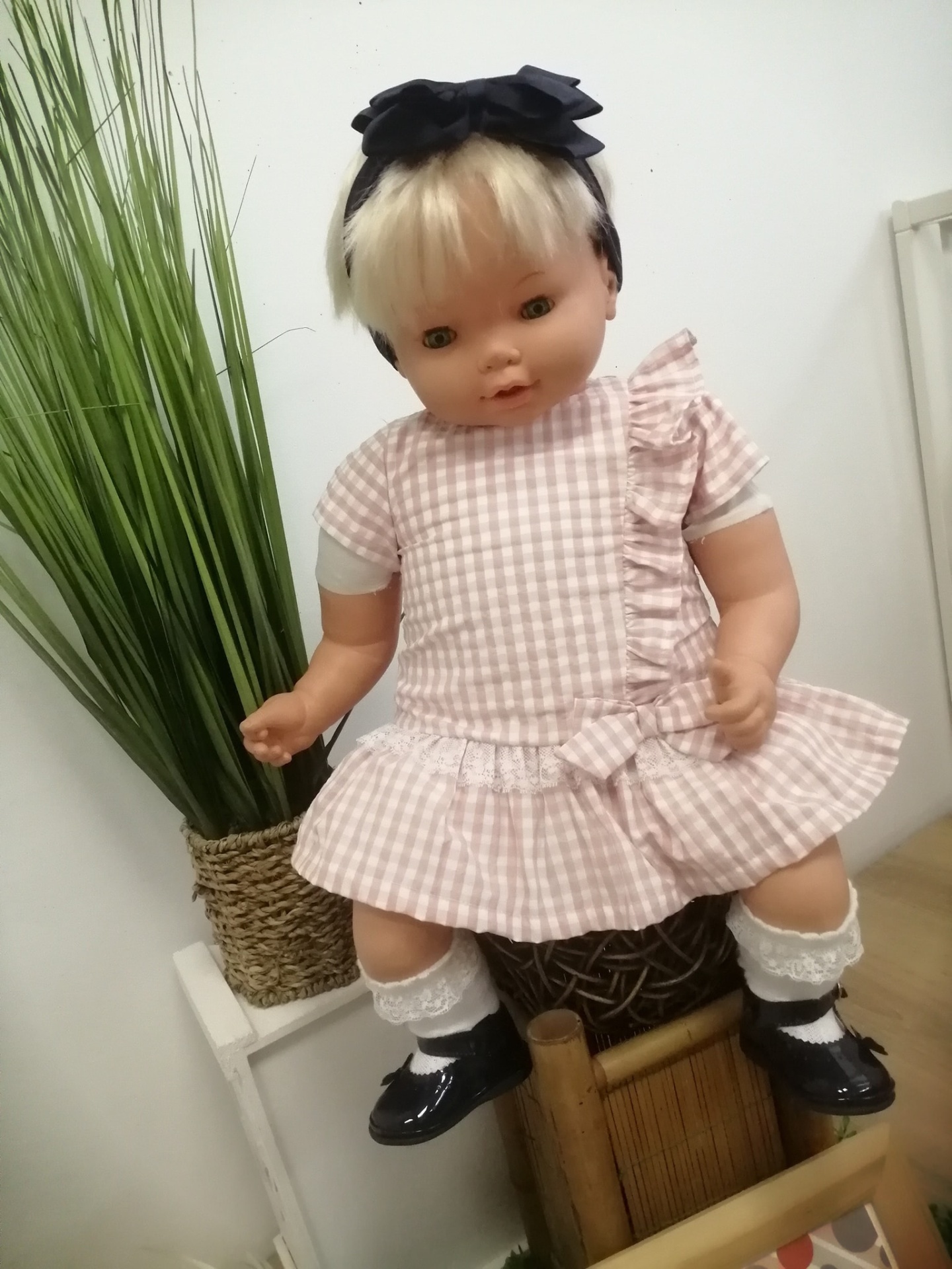 Boneca infantil com vestido xadrez rosa e branco, meias de renda, sapatos pretos e faixa de cabelo preta sentada numa cadeira