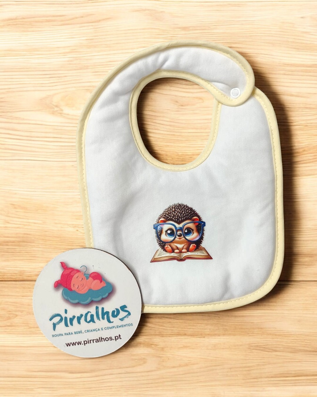 Babete branco com ouriço e etiqueta da Pirralhos sobre fundo de madeira