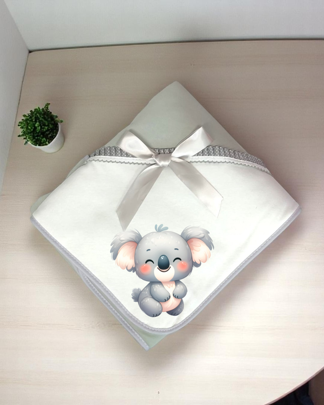 Manta de bebé branca com fita e desenho de coala, sobre mesa de madeira clara com planta em vaso pequeno