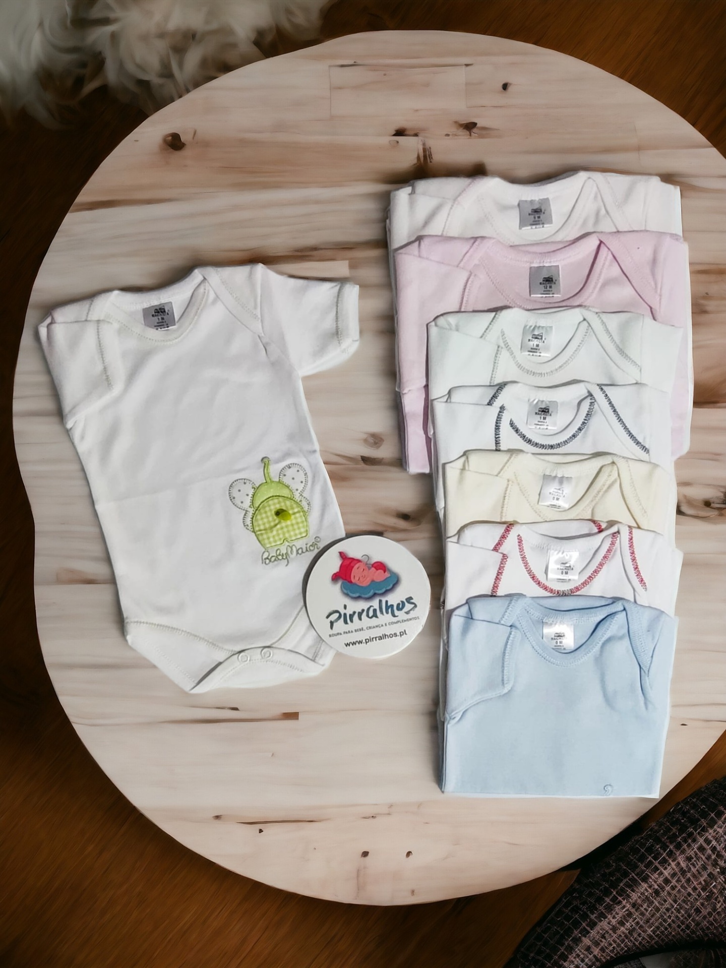Sete bodies de bebé de várias cores e bordados sobre mesa de madeira com cartão redondo 'Pirralhos'.