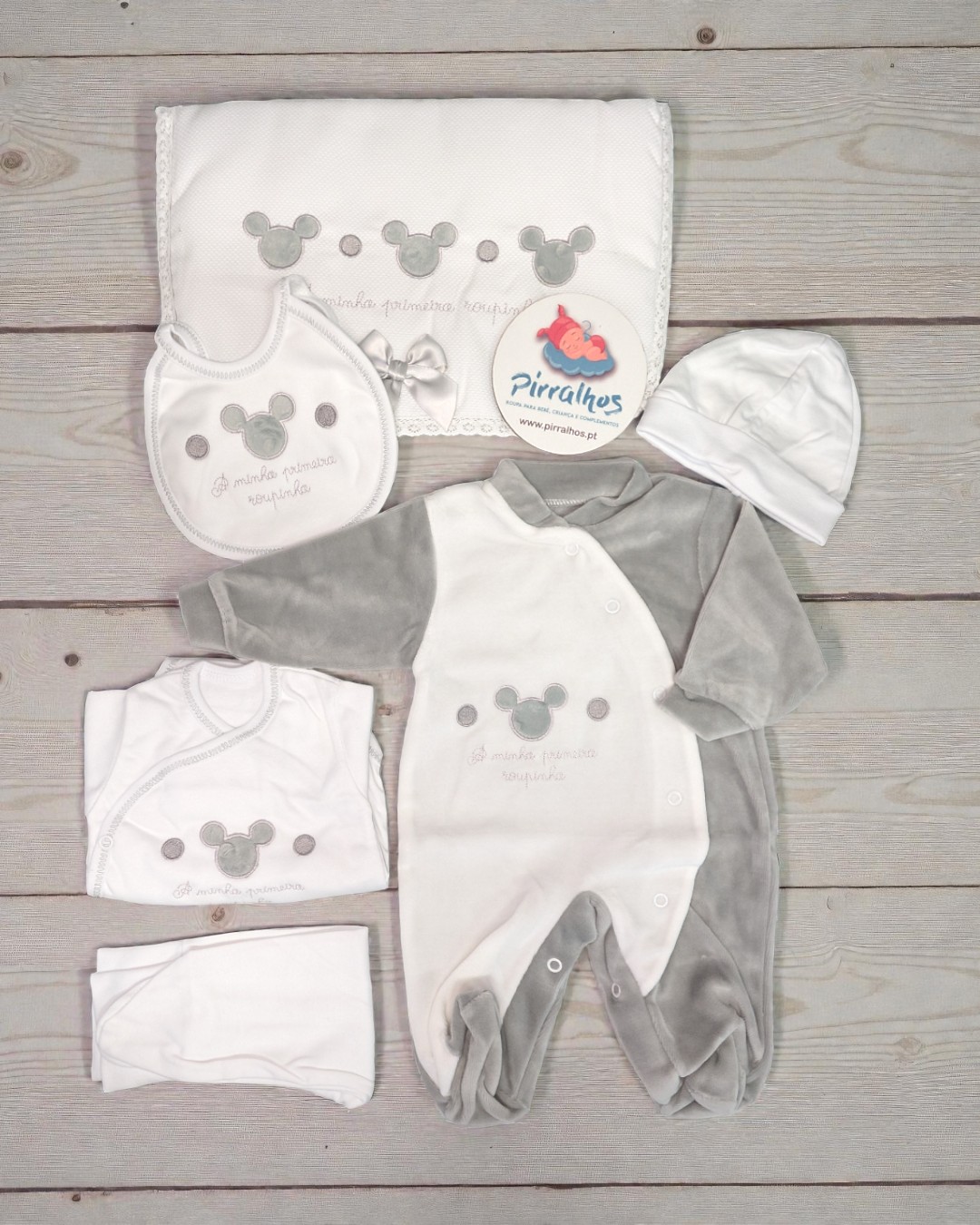 Conjunto de roupa de bebé branco e cinzento com estampas de ratinhos e texto A minha primeira roupinha, sobre piso de madeira.