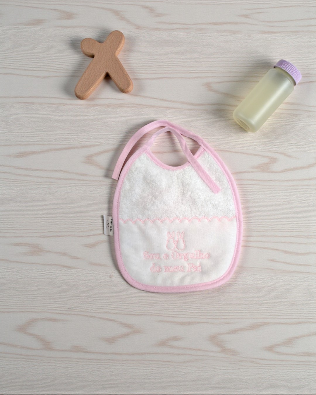 Babete de bebé branco e rosa com bordado, brinquedo de madeira e frasco plástico sobre superfície de madeira clara