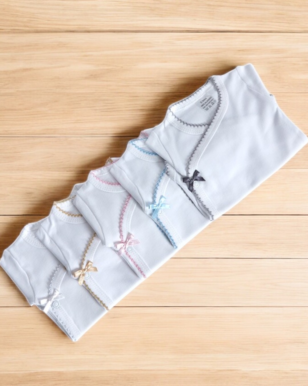 Bodies de bebé brancos com bordas coloridas e laços pequenos sobre superfície de madeira clara.