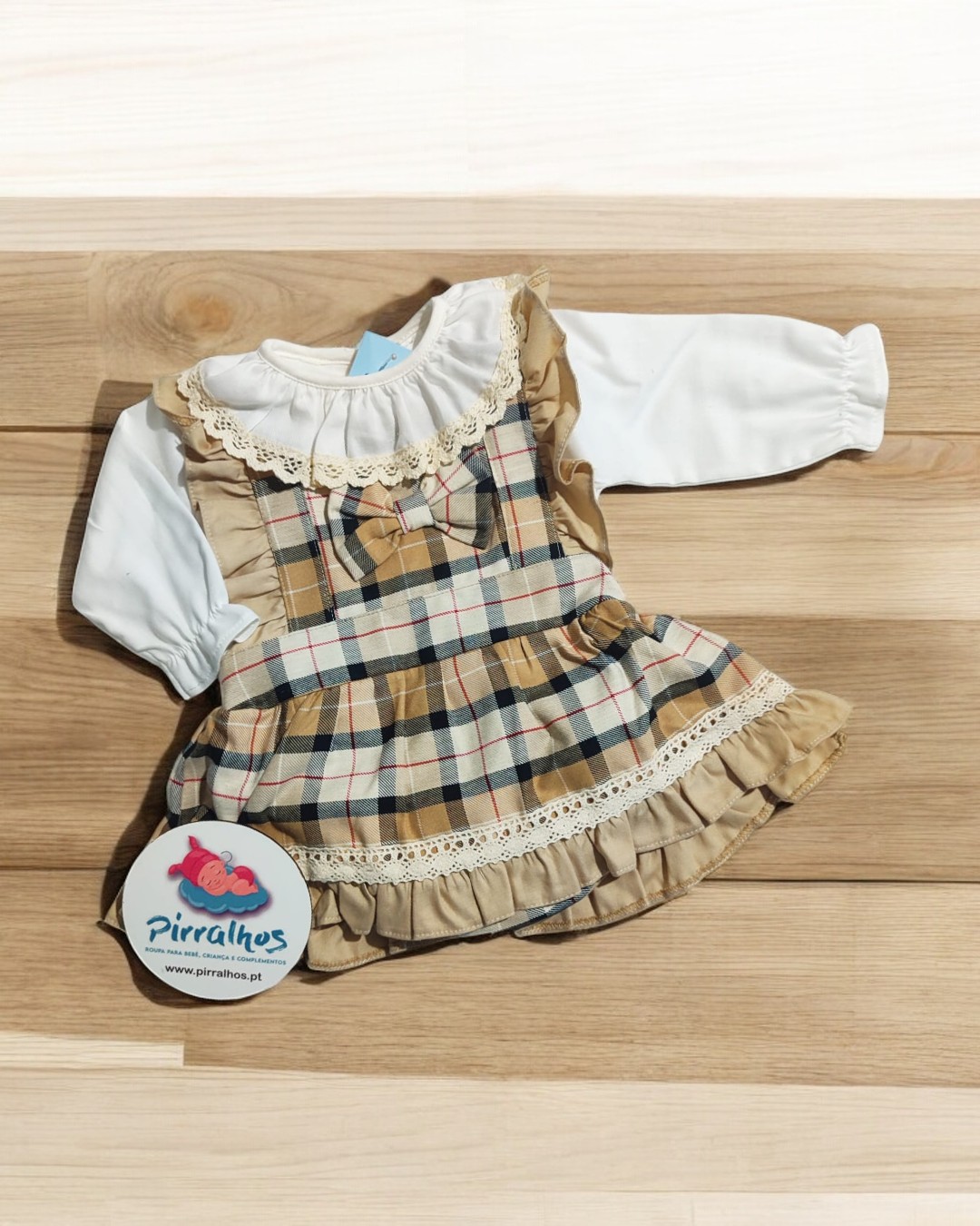Vestido infantil xadrez com detalhes rendados e calças brancas sobre mesa de madeira clara