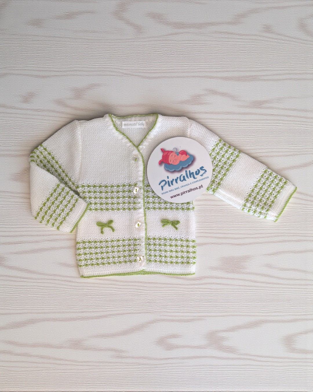 Camisola de bebé branca com padrões verdes e botões, sobre uma superfície de madeira clara.