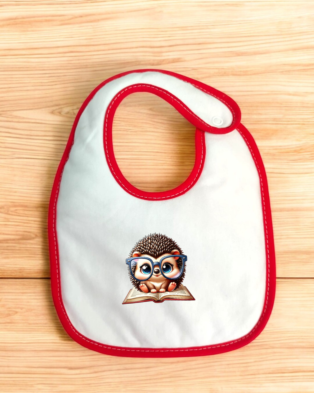 Babete branco com borda vermelha e estampa de ouriço com óculos azuis sobre um livro, colocado sobre superfície de madeira clara