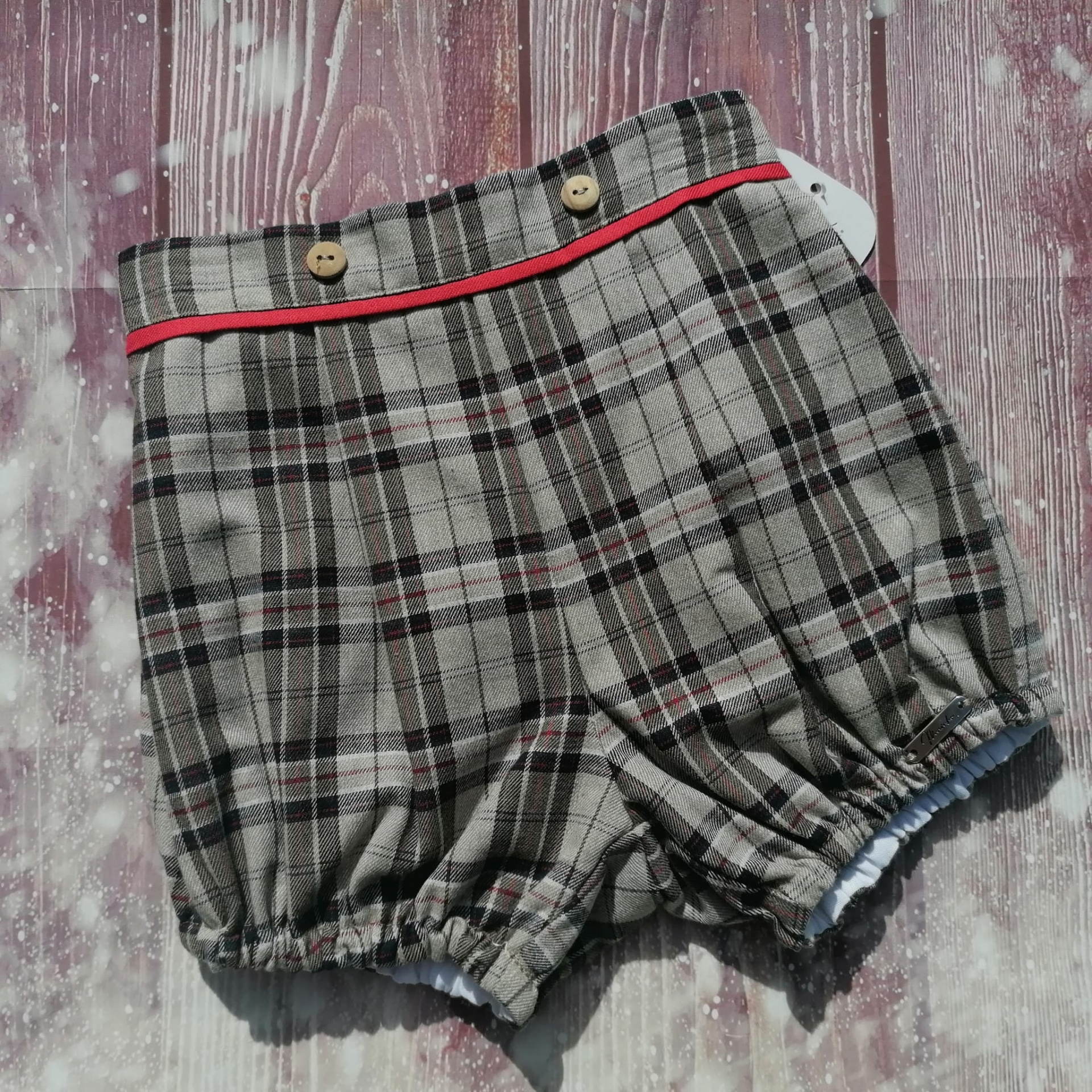 Calção infantil xadrez cinza com detalhes vermelhos e botões em fundo de madeira vinho
