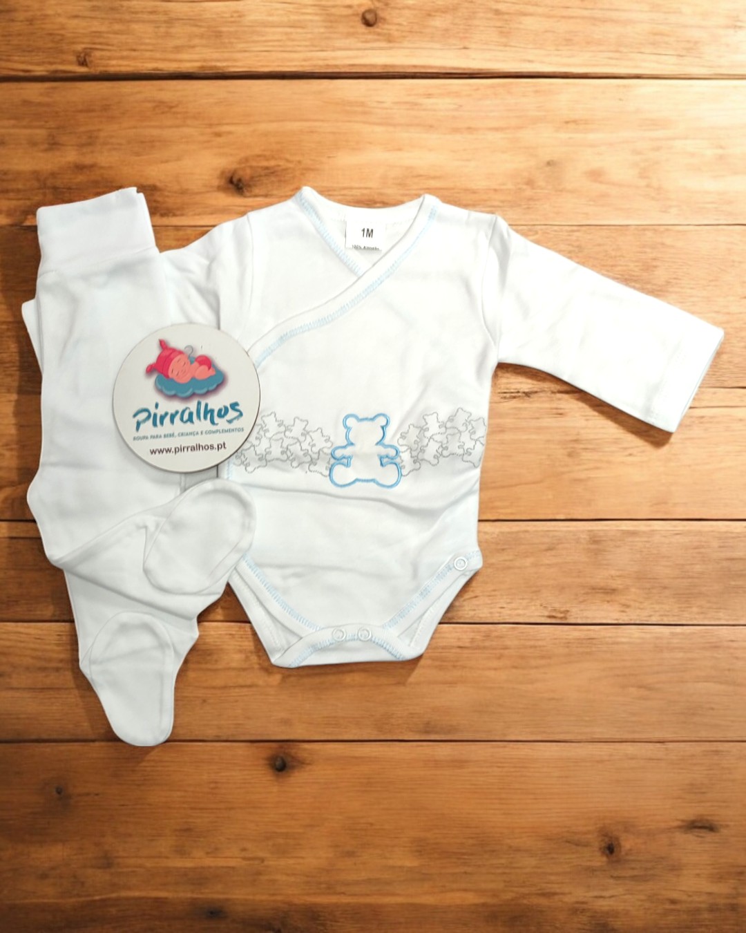Conjunto roupa bebé branco com ursinho azul e calças com pé, sobre madeira clara