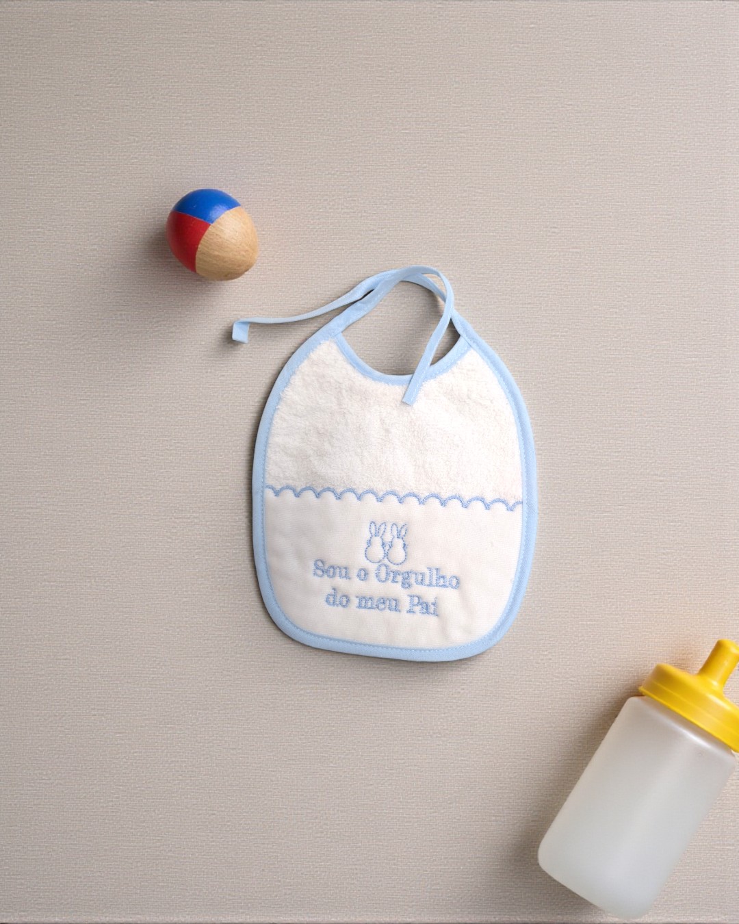 Babete branco com bordado azul, brinquedo colorido e biberão com tampa amarela.