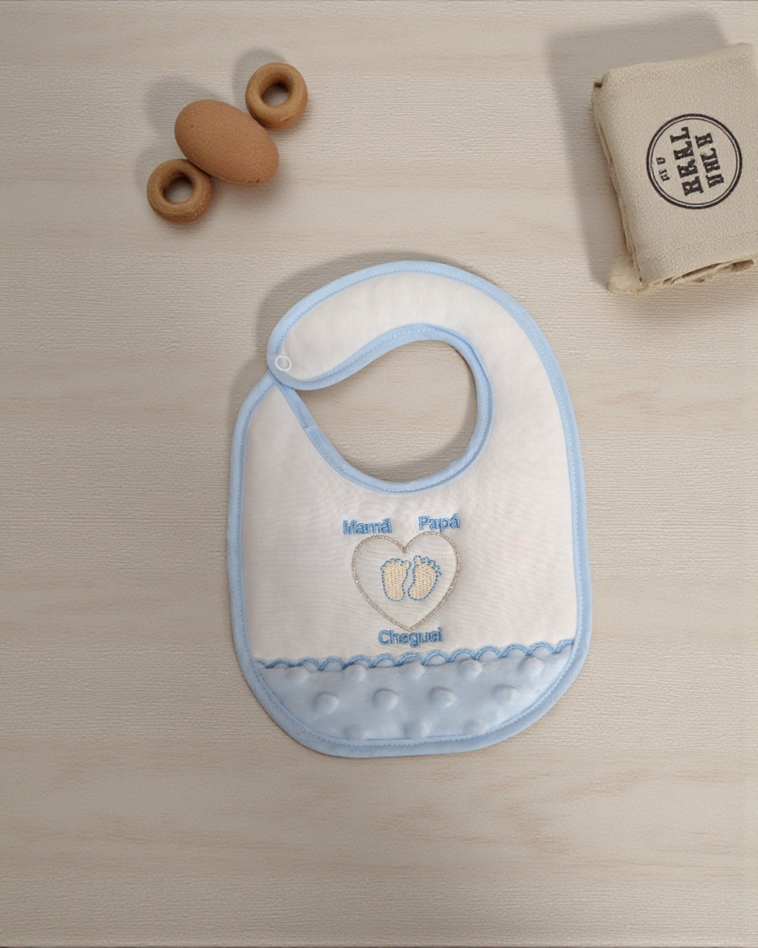 Babete branco com bordas azuis e bordado com texto 'Mamã Papá Cheguei' e pegadas azuis.