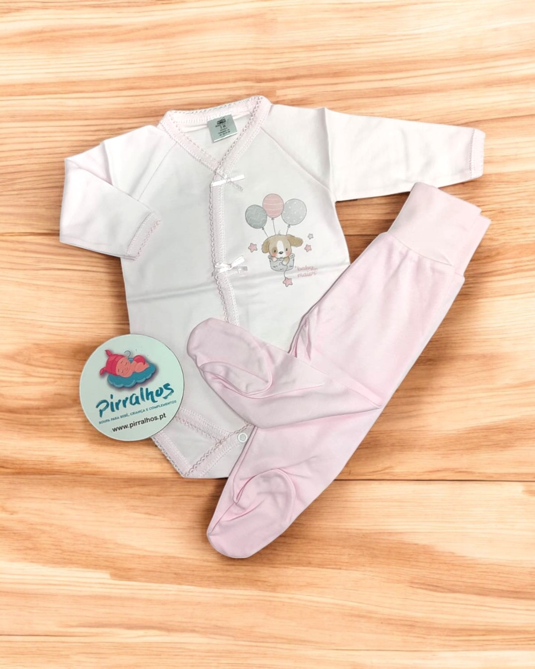 Conjunto de roupa de bebé branco e rosa com desenho de cão e balões, sobre fundo de madeira