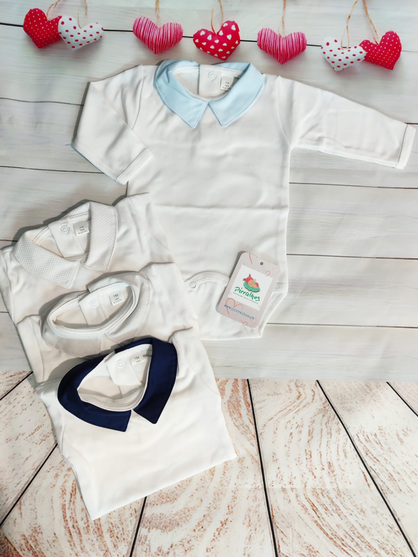 Bodies de bebé branco com golas coloridas e decoração de corações em tecido vermelho e branco ao fundo