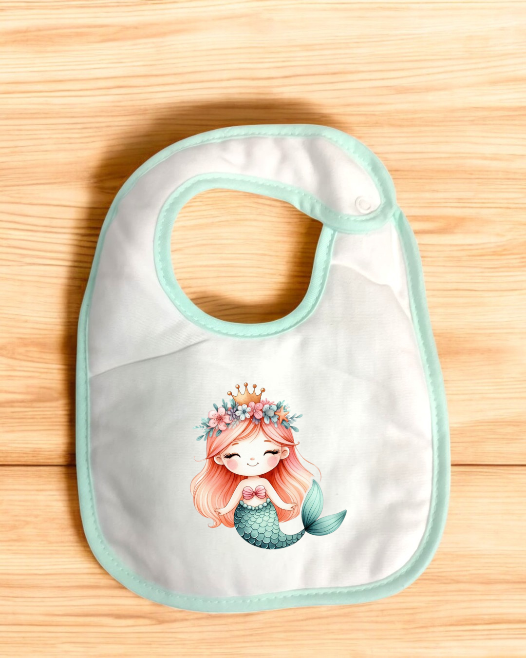 Babete branco com bordas verdes e sereia desenhada, colocado sobre madeira clara.