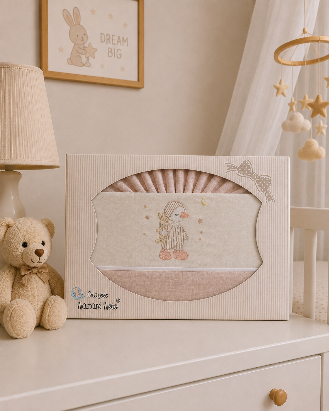 Conjunto de roupa de bebé cor de rosa embalado numa caixa decorada, sobre uma cómoda, ao lado de urso de peluche e lâmpada