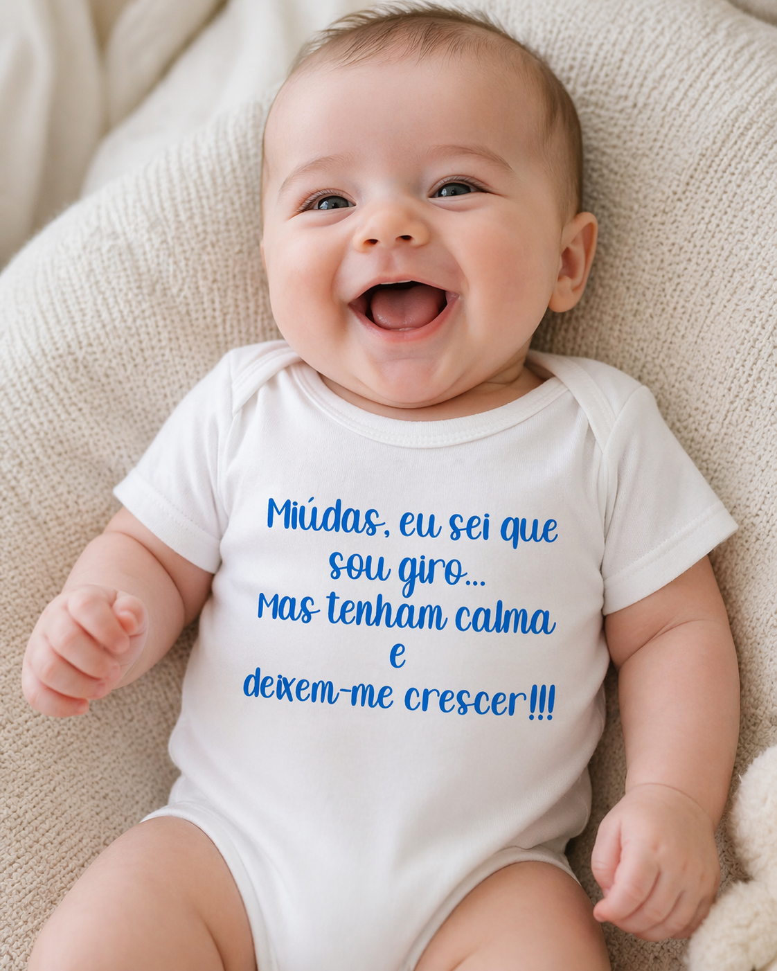 Body de bebé branco com texto azul sentado num tecido bege texturizado