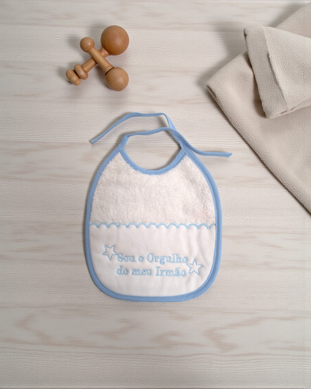 Babete branco com detalhes azuis e bordado texto em português sobre superfície de madeira clara