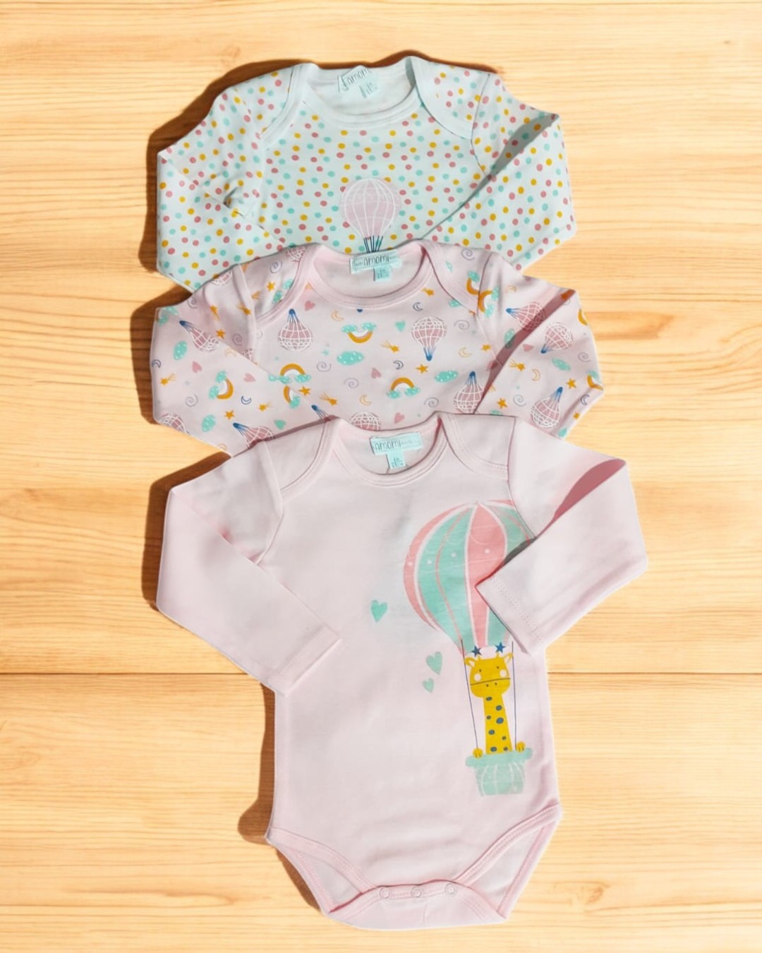 Três bodies bebés coloridos com padrões e ilustrações, mangas compridas, sobre tábua de madeira.