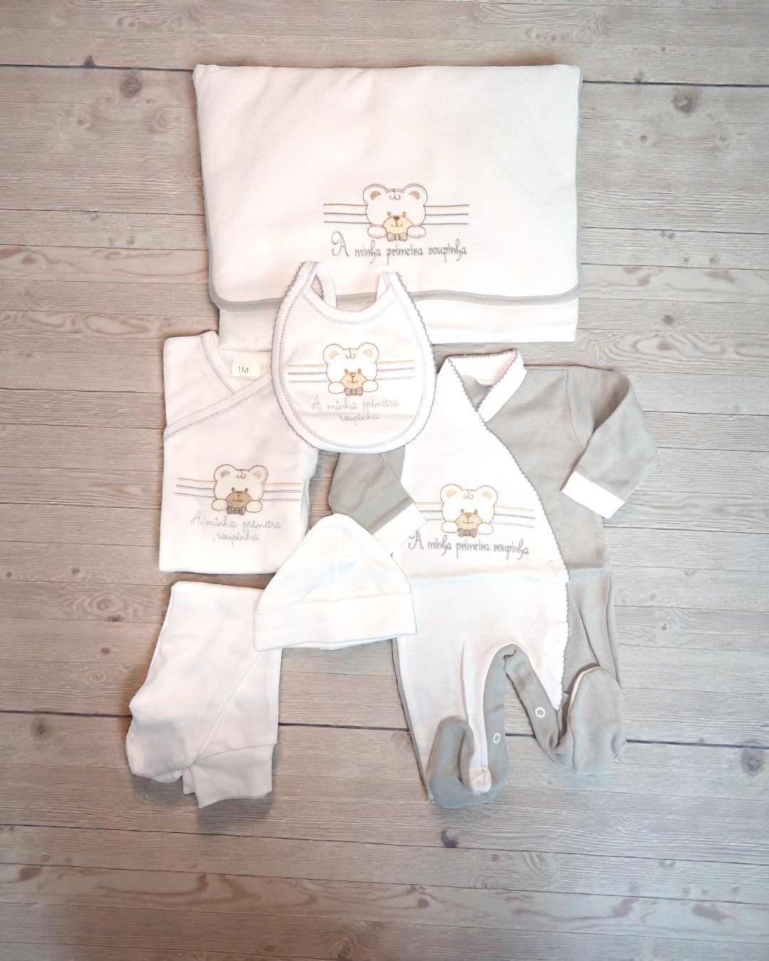 Conjunto de roupa e acessórios para bebé branco e cinza com ursinho bordado sobre madeira clara